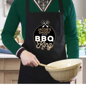 Personalised BBQ Apron for the BBQ King: A man in a green jumper wears a custom black cotton apron while holding a mixing bowl. The apron features a gold and white circular design with the text 'JACOB'S DADDY IS THE BBQ KING.' Set in a bright, rustic kitchen.