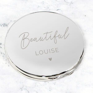 A studio-lit, high-angle close-up of a round, silver compact mirror resting on a white marble background. The mirror is engraved with the word "Beautiful" in elegant script, followed by the name "LOUISE" and a small heart icon.