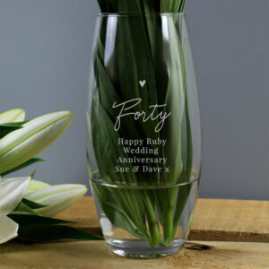 A clear, bullet-shaped glass vase on a wooden surface, filled with fresh green foliage. The glass is engraved with a small heart icon, the word "Forty," and the text "Happy Ruby Wedding Anniversary" along with "Sue & Dave x".