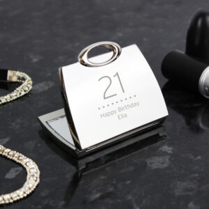 A high-angle studio shot of a shiny, silver nickel-plated handbag-shaped compact mirror, open on a dark marble surface. The lid is engraved with the number "21" and the message "Happy Birthday Ella." A sparkling rhinestone bracelet sits to the left, and a tube of lipstick is positioned to the right.
