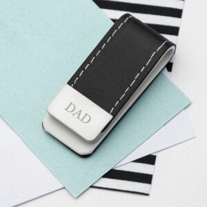 A close-up studio shot of a black PU leather money clip with cream stitching, resting on a background of blue and white paper. The stainless steel clip is engraved with the initials "DAD" in a traditional, elegant Classic Serif font.