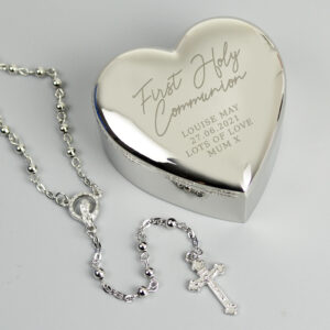 Personalised silver heart-shaped nickel-plated trinket box engraved with 'First Holy Communion' next to a silver rosary bead necklace, presented on a light grey background.