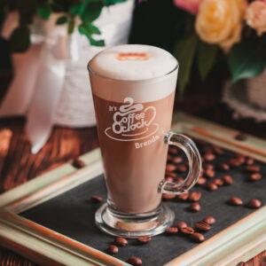 An elegant latte glass filled with coffee and foam, engraved with "It's Coffee O'Clock" and the name "Brenda," sitting on a wooden frame surrounded by coffee beans.