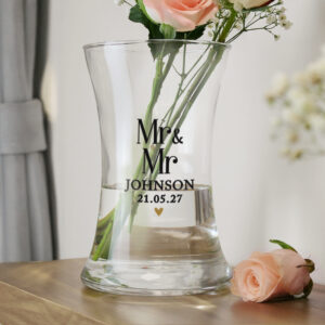 A clear, waisted glass vase sitting on a wooden surface next to a single pink rose. The vase is engraved with the text "Mr & Mr", "JOHNSON", and the date "21.05.27" with a small gold heart beneath.
