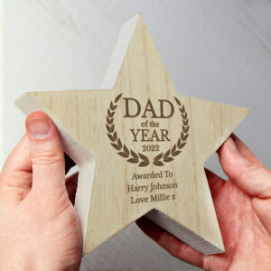 Personalised Dad Of The Year Star: A chunky, free-standing wooden star award being held between two hands. The star features a natural wood grain front with an engraved laurel wreath and the text "DAD of the YEAR 2022," followed by the personalised message "Awarded To Harry Johnson Love Millie x."