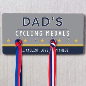 Personalised Dad's Achievement Medal Hooks: A grey and navy rectangular personalised medal rack hanging on a wall, featuring the text "DAD'S CYCLING MEDALS," five yellow stars, and a personalised message below reading "OUR #1 CYCLIST, LOVE CHLOE." Two medals with red, white, and blue ribbons are shown hanging from the hooks.