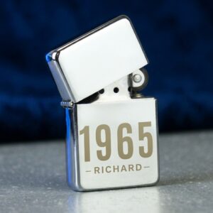 A polished silver flip-top lighter standing upright, engraved with the year "1965" and the name "RICHARD" centered below it, set against a dark blue background on a speckled surface.