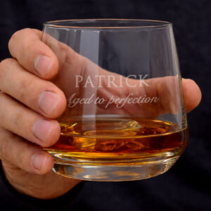 Personalised Definition Whisky Tumbler: A hand holding a heavy-based glass tumbler filled with whisky. The engraving features the name "PATRICK" in block capitals with the phrase "Aged to perfection" in an elegant script beneath it.