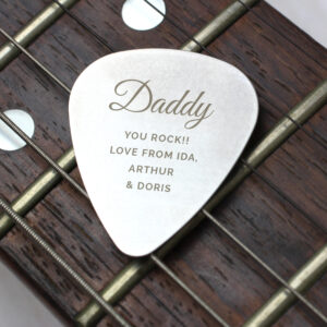 Personalised Father's Day Plectrum: A stainless steel guitar pick resting on the dark wood and strings of a guitar fretboard. The plectrum is engraved with "Daddy You Rock!! Love from Ida, Arthur & Doris."