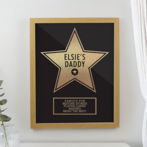 Personalised Father's Day Star Award Frame: A gold star award design in a light wood frame, hanging on a white wall. The star features the text "Elsie's Daddy" and a plaque listing things he is famous for, like "Bedtime Stories" and "Napping."