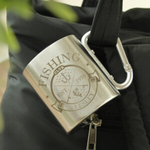 Personalised Fishing Club Mug: A silver stainless steel mug with a carabiner handle clipped to a black bag. The mug is engraved with a "Fishing Club" crest featuring a fish and hooks, personalised with the name "LESLEY" and the year "1956."