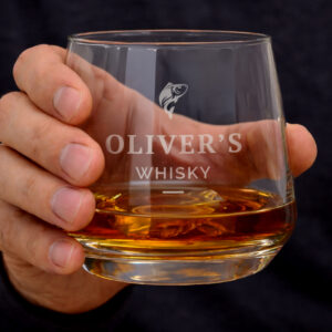 Personalised Fishing Whisky Glass: A man's hand holding a heavy-based glass tumbler filled with amber whisky. The glass is engraved with a small jumping fish motif, the name "OLIVER'S," and the word "WHISKY."