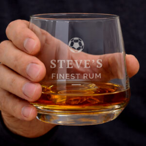 Personalised Football Tumbler: A close-up of a hand holding a weighted glass tumbler filled with amber rum. The glass is engraved with a football icon, the name "STEVE'S," and "FINEST RUM."