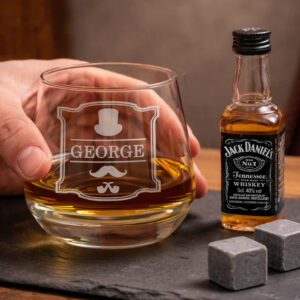 Personalised Gentlemen Tumbler And Whisky Miniature Set: A hand holding a whisky tumbler engraved with the name "GEORGE" and a gentleman-themed motif, sitting on a slate coaster next to a miniature bottle of Jack Daniel's and two whisky cooling stones.