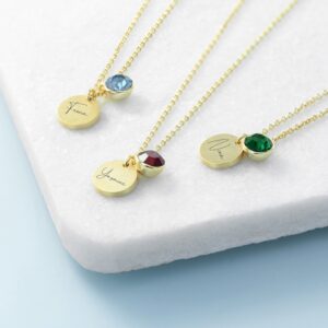 A close-up photograph displays three delicate gold chain necklaces with small charms resting on a white, rough-textured marble surface with a subtle blue gradient background. Each necklace features two pendants: one circular, matte gold disc disc and one circular, faceted birthstone crystal. The necklace on the far left features a light blue crystal (potentially aquamarine) and a gold disc engraved with the name 'Fiona' in an elegant script font. The necklace in the center holds a deep red crystal (potentially garnet or ruby) and a gold disc engraved with the name 'Yasmine' in a script font. The necklace on the right shows a vibrant green crystal (potentially emerald) and a gold disc engraved with the name 'Nina' in a script font. The three necklaces are draped parallel to each other.