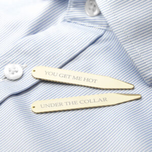 A close-up studio shot of two 18K gold-plated collar stiffeners resting on a crisp, light blue and white pinstriped men's dress shirt. The top stiffener is engraved with the text "YOU GET ME HOT" and the one below it reads "UNDER THE COLLAR" in a sophisticated, elegant Classic font.