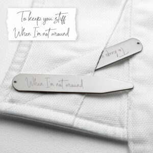 A close-up photograph of a pair of silver rhodium-plated collar stiffeners resting on a crisp white dress shirt. One stiffener is inserted into the collar fold, revealing the engraved handwriting text "When I'm not around." Above the shirt, a strip of white paper displays the original handwritten note: "To keep you stiff / When I'm not around".
