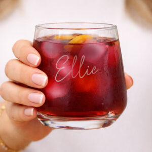 Personalised Name Glass Tumbler: A close-up of a hand holding a weighted glass tumbler filled with a dark red drink and ice. The name "Ellie" is elegantly engraved on the front in a flowing script font.