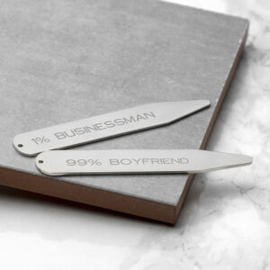 A close-up photograph of two silver rhodium-plated collar stiffeners resting on a grey stone tile. The top stiffener is engraved with the text "1% BUSINESSMAN" and the bottom one with "99% BOYFRIEND" in a clean, modern Sans Serif font. The surface has a marble-like texture in the corner.