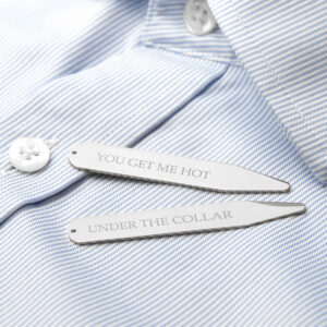 A close-up studio shot of two silver rhodium-plated collar stiffeners resting on a crisp, light blue and white pinstriped men's dress shirt. The stiffener at the top is engraved with the text "YOU GET ME HOT" and the one below it reads "UNDER THE COLLAR". The shirt has a white button visible in the background.