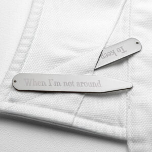 A close-up photograph of a pair of high-shine silver rhodium-plated collar stiffeners resting on a textured white dress shirt. The white shirt material is folded over, and one stiffener is tucked slightly into the fold, mimicking how it is inserted into a shirt collar. This stiffener clearly reveals the text "When I'm not around" engraved in an elegant, classic Serif font. The other stiffener is mostly hidden within the fold.