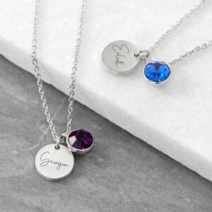 Close-up of two silver necklaces resting on a white stone background; each features a round, personalised silver disc engraved with names "Georgia" and "Eilidh" accompanied by a single purple and blue birthstone crystal charm.