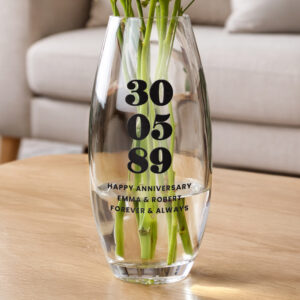 A clear, bullet-shaped glass vase resting on a wooden table. The vase features an engraved date "30 05 89" in large, bold numbers, followed by the text "HAPPY ANNIVERSARY," "EMMA & ROBERT," and "FOREVER & ALWAYS".