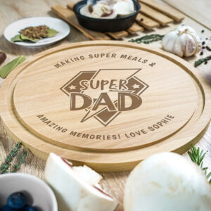 Personalised Super Dad Chopping Board: A round wooden chopping board engraved with a central "Super Dad" logo and a personalised message around the edge that reads "Making Super Meals & Amazing Memories! Love Sophie," surrounded by fresh cooking ingredients like garlic, mushrooms, and herbs.