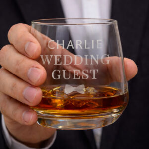 Personalised Wedding Whisky Glass: A heavy-bottomed whisky tumbler held by a man in a suit. The glass is engraved with "Charlie Wedding Guest" and a small bow tie motif, filled with amber-coloured spirits.