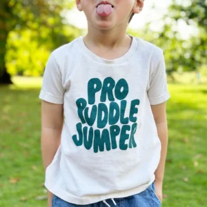 A young boy pulling a funny face, wearing a natural-colored organic cotton t-shirt with the text "Pro Puddle Jumper" in teal, standing in a lush green park setting.