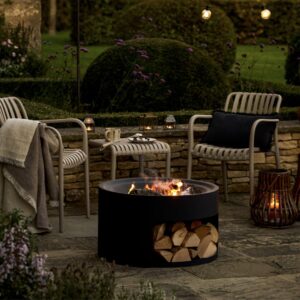 Matte black round steel fire pit with a log store base and glowing fire on a stone patio with garden seating.