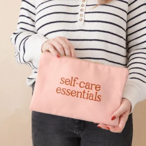 A person wearing a striped long-sleeved top holding a blush pink canvas pouch with the words "self-care essentials" printed in orange, with their hand opening the metal zip.
