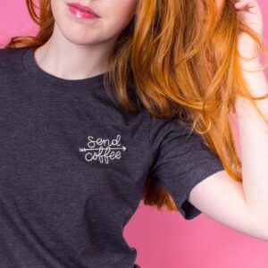 A person with long red hair wearing a dark heather grey unisex t-shirt, embroidered with the text 'send coffee' and an arrow design in off-white thread.