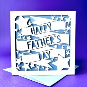 Stars Papercut Father's Day Card: A square white Father’s Day card featuring a laser-cut design of stars and flowing banners with the words "HAPPY FATHER'S DAY" stencilled out. The design reveals a blue background and sits atop a matching light blue envelope against a purple backdrop.