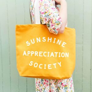 A person wearing a floral dress carrying a vibrant yellow canvas tote bag that reads "SUNSHINE APPRECIATION SOCIETY" in bold off-white lettering, against a matching green wooden background.