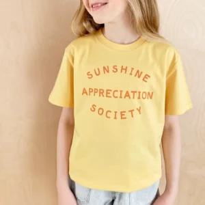 A child modeling a honeycomb yellow kid’s t-shirt featuring the text "Sunshine Appreciation Society" printed in an orange-yellow font, set against a simple wooden background.
