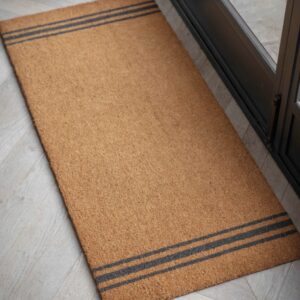 A high-angle, close-up shot of an extra-long coir doormat with a classic triple-stripe design, placed on a light wood floor near a door frame.