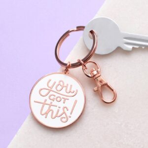 A white enamel keyring with copper trim featuring the handwritten text You Got This. The keyring has a split ring and a lobster clasp, resting on a soft-textured background.