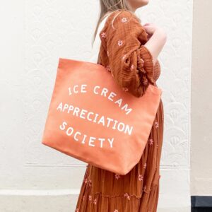 A person wearing a burnt orange floral dress holding a peach-colored canvas tote bag that reads 'Ice Cream Appreciation Society' in bold white text, standing against a textured white wall.