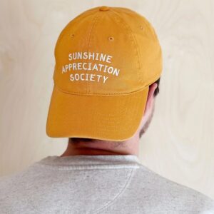 Back view of a yellow cotton baseball cap with white embroidered text that reads Sunshine Appreciation Society, worn by a person on a neutral background