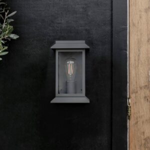 Modern carbon steel lantern wall light with a matte finish and glass framing, mounted on a dark exterior wall next to a natural wood door frame.