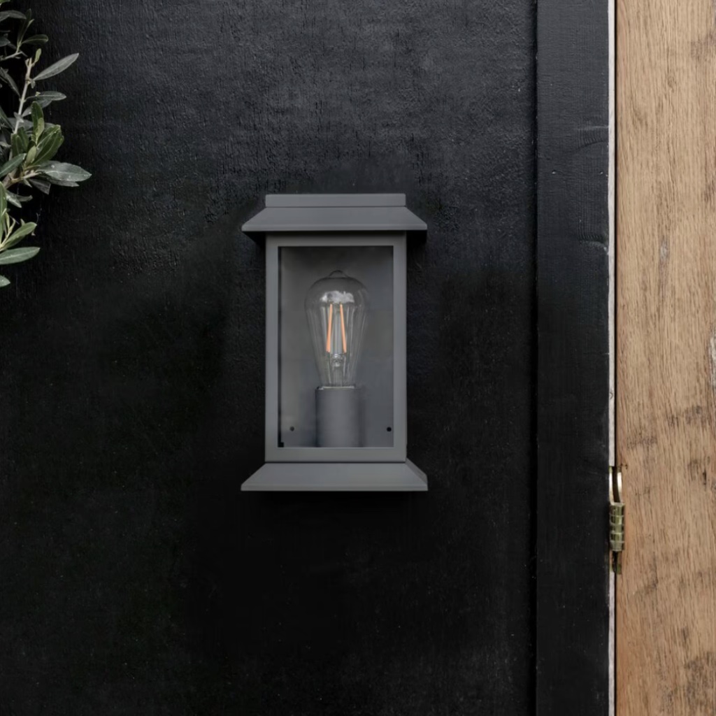 Modern carbon steel lantern wall light with a matte finish and glass framing, mounted on a dark exterior wall next to a natural wood door frame.
