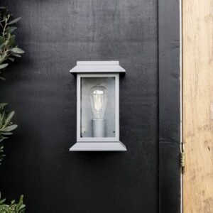 Modern charcoal steel lantern wall light with a matte finish and clear glass frame, mounted on a black wooden exterior wall next to a light oak door.