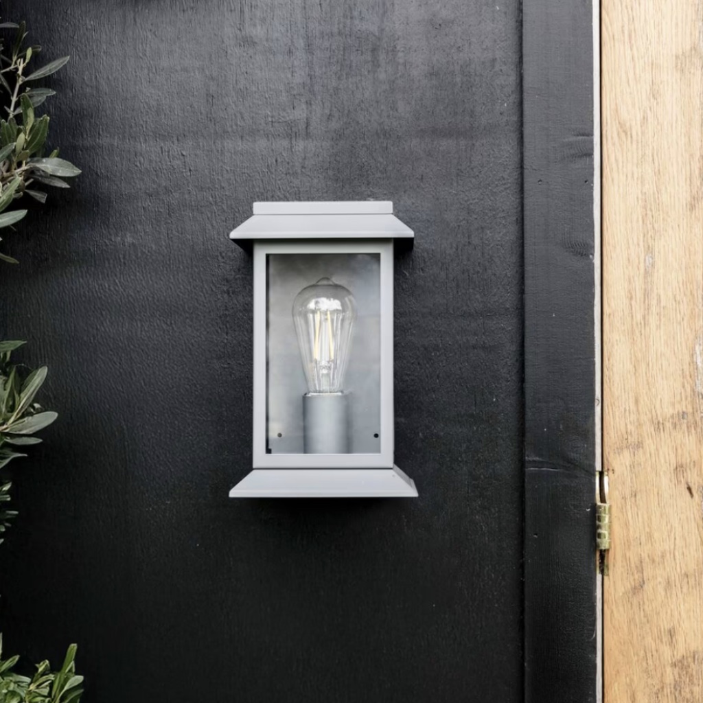 Modern charcoal steel lantern wall light with a matte finish and clear glass frame, mounted on a black wooden exterior wall next to a light oak door.