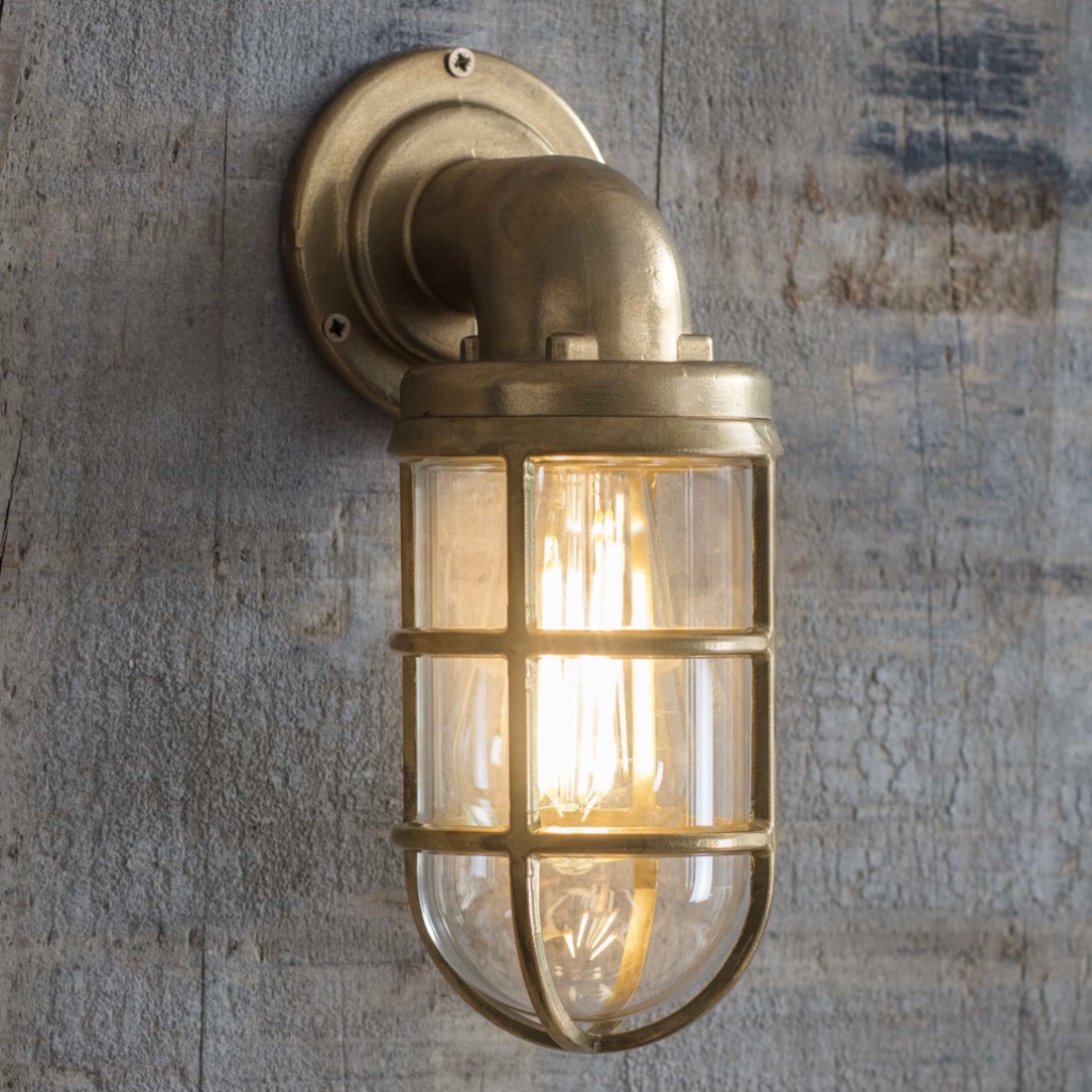Nautical raw cast brass down light with an industrial cage design and lit filament bulb, mounted on a weathered wood wall.