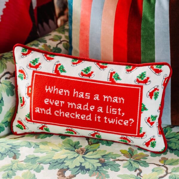 Check It Twice hand-stitched needlepoint Christmas cushion resting on an armchair alongside colourful velvet pillows