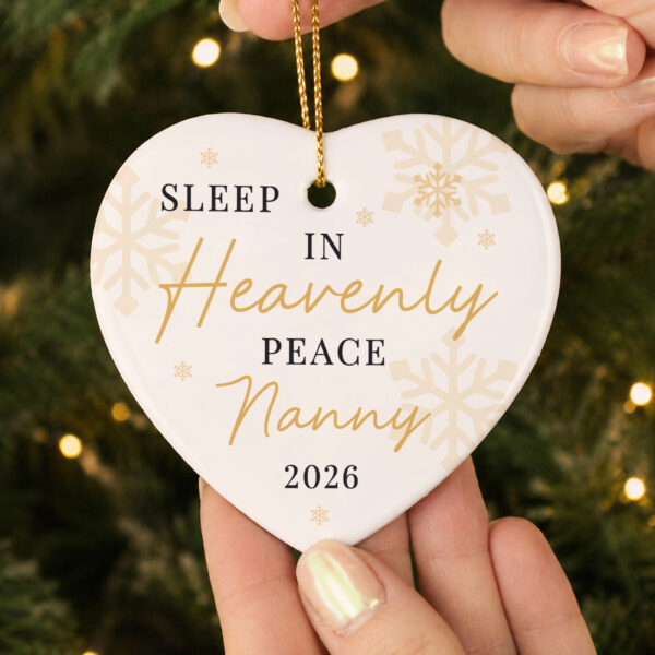 Hands holding a white ceramic heart Christmas decoration personalised with the name Nanny and year 2026 beneath Sleep In Heavenly Peace text.