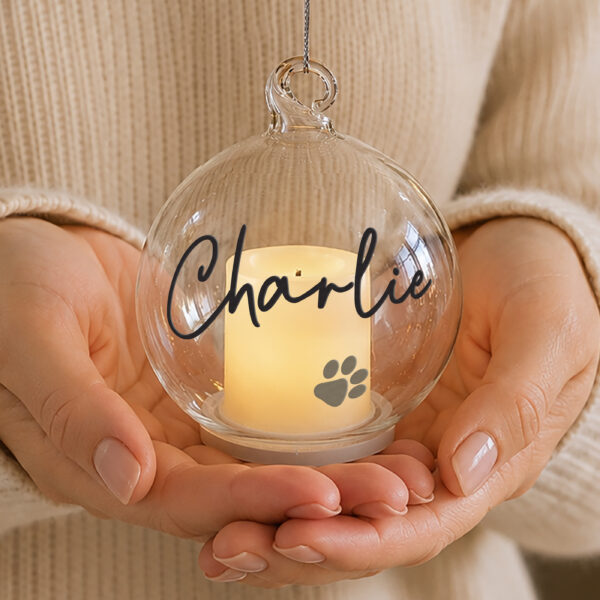 Hands gently holding a clear glass LED candle bauble personalised with the name Charlie and a pawprint.