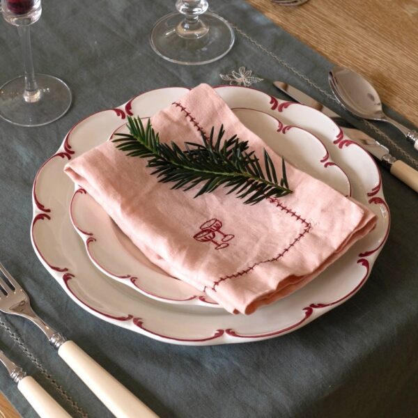 Plaster pink embroidered linen napkin folded on a scalloped plate over a dark green linen tablecloth with place setting